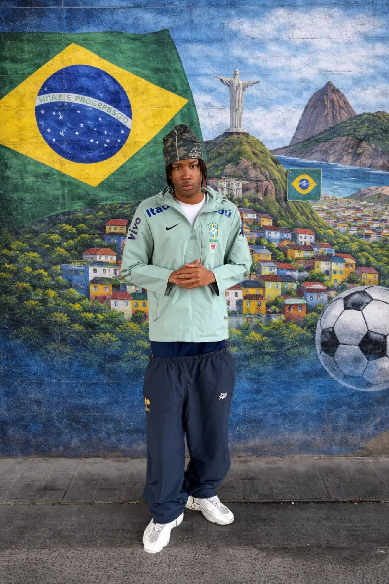 Client Drip Seleção en veste imperméable verte devant une fresque du drapeau brésilien et du Corcovado